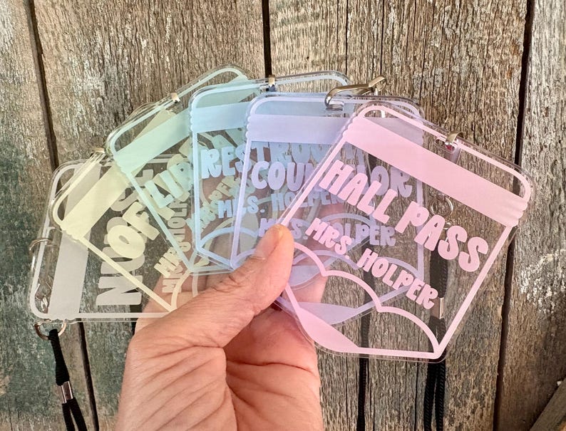 May include: A set of clear acrylic rectangular hall passes with rounded corners, each featuring pastel-colored text and a matching lanyard. The passes display various labels including Hall Pass, Library, and Restroom, along with the name Mrs. Holper.
