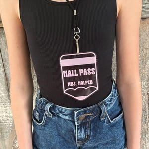 May include: A clear acrylic hall pass shaped like a pencil with light pink accents. The text HALL PASS is printed in a bold font above the name MRS. HOLPER. The pass hangs from a black lanyard with a silver metal clip attached to the top.