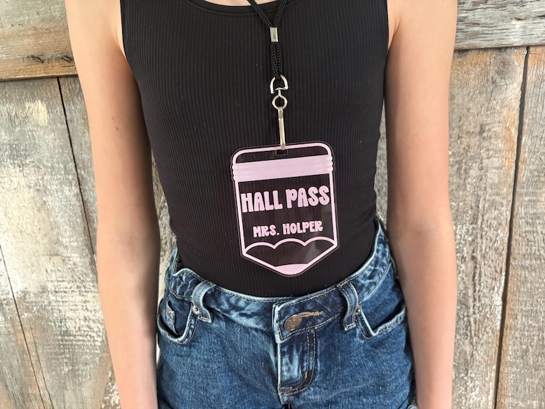 May include: A clear acrylic hall pass shaped like a pencil with light pink accents. The text HALL PASS is printed in a bold font above the name MRS. HOLPER. The pass hangs from a black lanyard with a silver metal clip attached to the top.