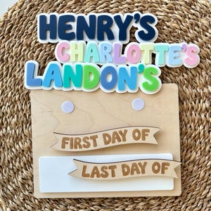 K&ouml;nnte beinhalten: Ein Holzschild mit drei gestapelten Namensausschnitten: Henry's in Dunkelblau, Charlotte's in Pastellfarben und Landon's in Blau und Gr&uuml;n. Darunter zeigt ein Holzbrett zwei ausgeschnittene Banner mit der Aufschrift First Day Of und Last Day Of.