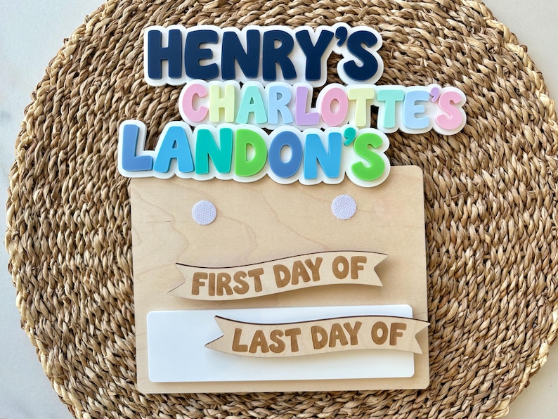 K&ouml;nnte beinhalten: Ein Holzschild mit drei gestapelten Namensausschnitten: Henry's in Dunkelblau, Charlotte's in Pastellfarben und Landon's in Blau und Gr&uuml;n. Darunter zeigt ein Holzbrett zwei ausgeschnittene Banner mit der Aufschrift First Day Of und Last Day Of.