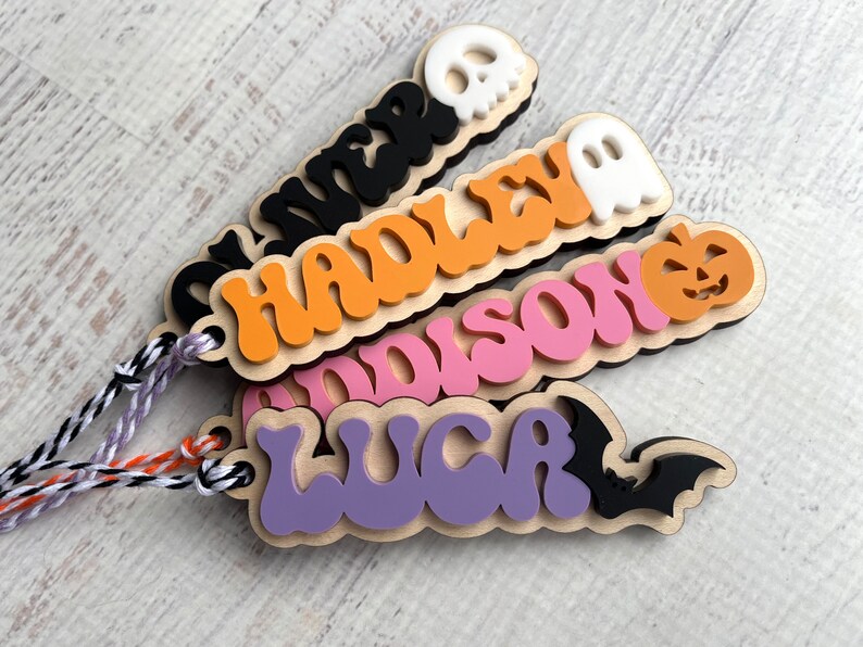 May include: Four wooden Halloween name tags with different colored backgrounds and Halloween-themed cutouts. The names are "Oliver", "Hadley", "Addison", and "Lucas".