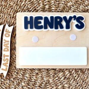 K&ouml;nnte beinhalten: Ein rechteckiges Holzschild mit dem dunkelblauen, erhabenen Schriftzug HENRY'S, zwei wei&szlig;en, runden Klettverschl&uuml;ssen und einer wei&szlig;en, rechteckigen Whiteboard-Fl&auml;che. Zwei separate Holzbanner tragen die Aufschriften FIRST DAY OF und LAST DAY OF.