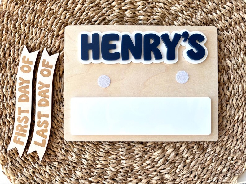 K&ouml;nnte beinhalten: Ein rechteckiges Holzschild mit dem dunkelblauen, erhabenen Schriftzug HENRY'S, zwei wei&szlig;en, runden Klettverschl&uuml;ssen und einer wei&szlig;en, rechteckigen Whiteboard-Fl&auml;che. Zwei separate Holzbanner tragen die Aufschriften FIRST DAY OF und LAST DAY OF.