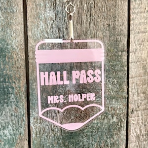 May include: A clear acrylic hall pass shaped like a pencil with light pink lettering that reads HALL PASS and MRS. HOLPER. The tag features a silver metal lobster clasp at the top for attachment, designed with a flat top and a pointed bottom.