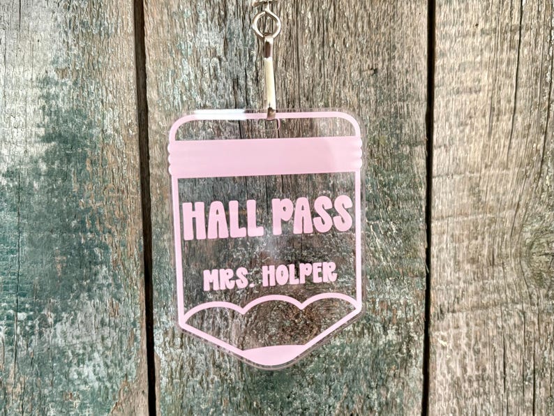 May include: A clear acrylic hall pass shaped like a pencil with light pink lettering that reads HALL PASS and MRS. HOLPER. The tag features a silver metal lobster clasp at the top for attachment, designed with a flat top and a pointed bottom.