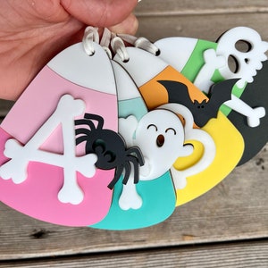 May include: Five Halloween-themed candy corn ornaments with different designs. The ornaments are pink, green, yellow, orange, and black. The designs include a spider, a ghost, a bat, a skull and crossbones, and the letter "A".
