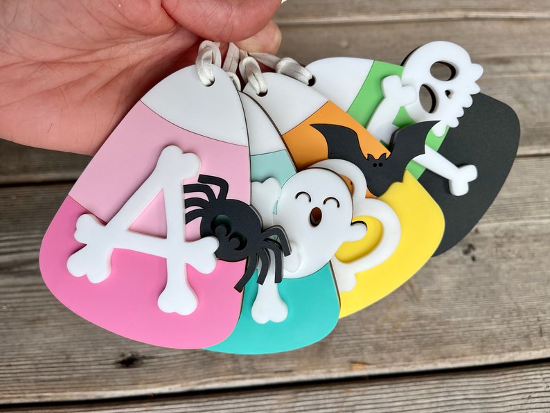 May include: Five Halloween-themed candy corn ornaments with different designs. The ornaments are pink, green, yellow, orange, and black. The designs include a spider, a ghost, a bat, a skull and crossbones, and the letter "A".