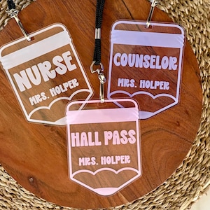 May include: Three clear acrylic badge holders shaped like pencils, each attached to a black lanyard. The badges feature white, lavender, and pink text reading NURSE, COUNSELOR, and HALL PASS, with the name MRS. HOLPER printed below each title.