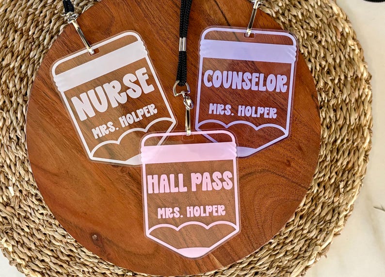 May include: Three clear acrylic badge holders shaped like pencils, each attached to a black lanyard. The badges feature white, lavender, and pink text reading NURSE, COUNSELOR, and HALL PASS, with the name MRS. HOLPER printed below each title.