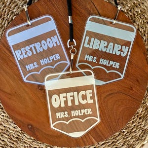 May include: Three clear acrylic pencil-shaped name tags with white lettering. Each tag features a top section, a central area with the text RESTROOM, LIBRARY, or OFFICE, and the name MRS. HOLPER printed below. The tags are attached to a black lanyard.