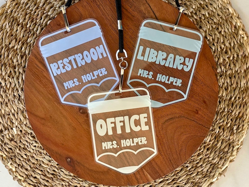May include: Three clear acrylic pencil-shaped name tags with white lettering. Each tag features a top section, a central area with the text RESTROOM, LIBRARY, or OFFICE, and the name MRS. HOLPER printed below. The tags are attached to a black lanyard.