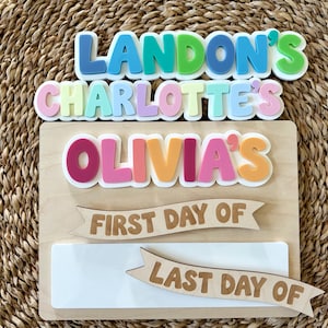 K&ouml;nnte beinhalten: Ein rechteckiges Holzschild mit bunten, erhabenen Acrylbuchstaben, die LANDON'S, CHARLOTTE'S und OLIVIA'S bilden. Unter den Namen befinden sich zwei h&ouml;lzerne Banner mit der Aufschrift FIRST DAY OF und LAST DAY OF sowie eine wei&szlig;e, abwischbare Fl&auml;che.