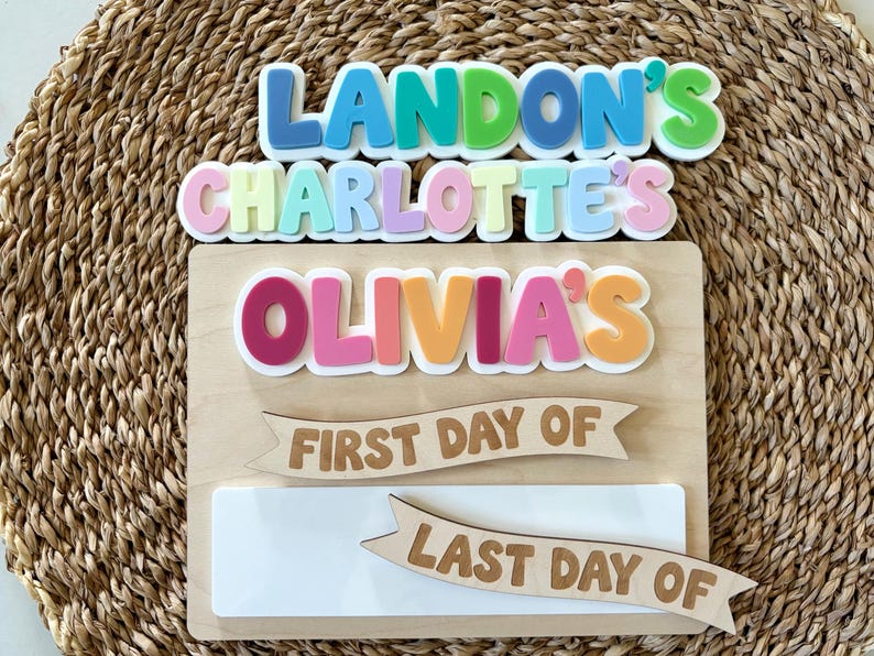 K&ouml;nnte beinhalten: Ein rechteckiges Holzschild mit bunten, erhabenen Acrylbuchstaben, die LANDON'S, CHARLOTTE'S und OLIVIA'S bilden. Unter den Namen befinden sich zwei h&ouml;lzerne Banner mit der Aufschrift FIRST DAY OF und LAST DAY OF sowie eine wei&szlig;e, abwischbare Fl&auml;che.