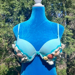 May include: A light blue bra decorated with seashells and beads. The bra is on a blue mannequin.