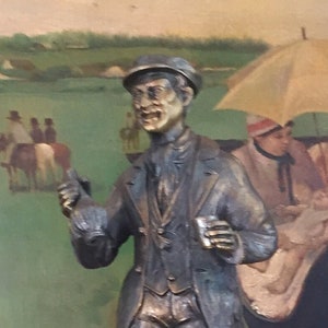 May include: A bronze-colored statue of a man in a suit and cap, holding a glass and gesturing. The statue is mounted on a stone base with a circular marble base. The background features a framed painting with a horse racing scene.