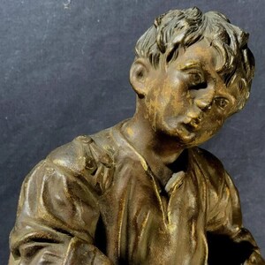 May include: A bronze sculpture of a young boy holding a shoe brush and a shoe. The boy is wearing a simple shirt and pants. The sculpture is detailed and realistic.