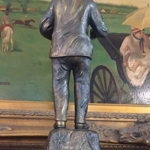 May include: A bronze statue of a man in a cap and jacket, viewed from the back. The statue stands on a stone base with engraved text, atop a marble-like circular base. The background features a painting of a horse race.