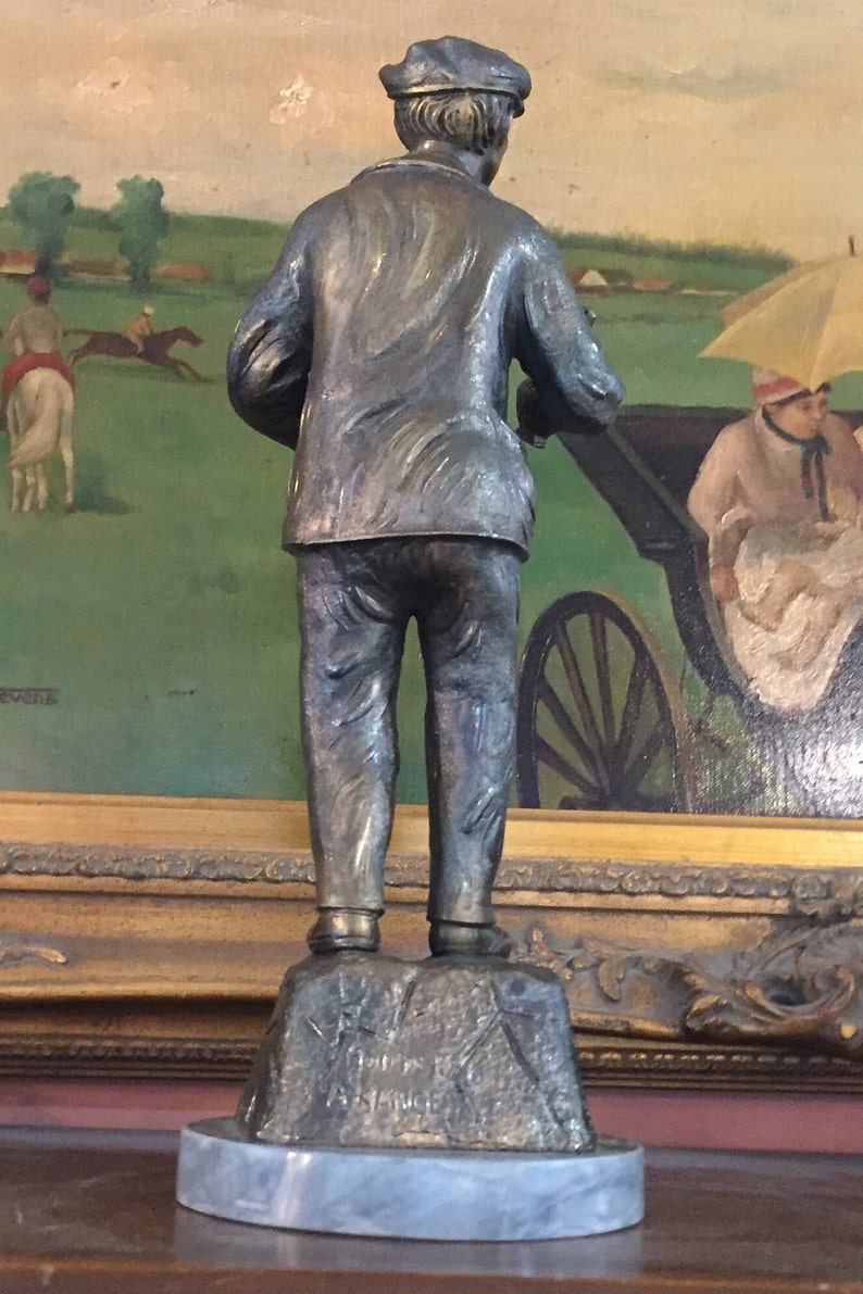 May include: A bronze statue of a man in a cap and jacket, viewed from the back. The statue stands on a stone base with engraved text, atop a marble-like circular base. The background features a painting of a horse race.