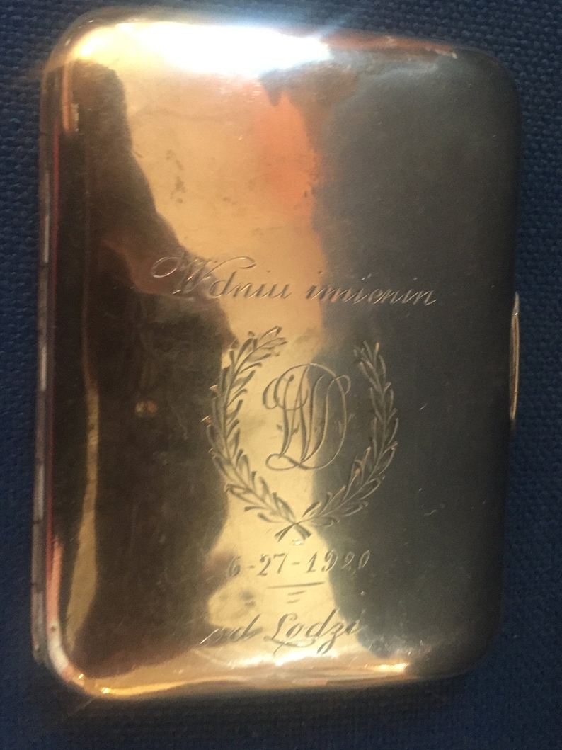 Op de afbeelding: Een gouden metalen sigarettenkoker met een gegraveerde inscriptie die "W dniu imienin" en "6-27-1920" en "od Lodzi" luidt.
