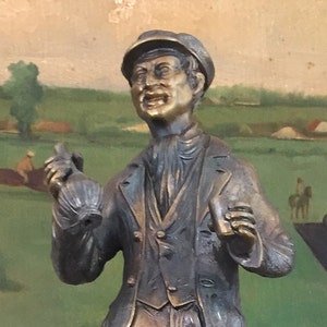 May include: A bronze statue of a man in a cap and coat, holding a bottle and a cup, with a painted backdrop of a horse race. The statue has a detailed, aged appearance, capturing a moment of excitement.