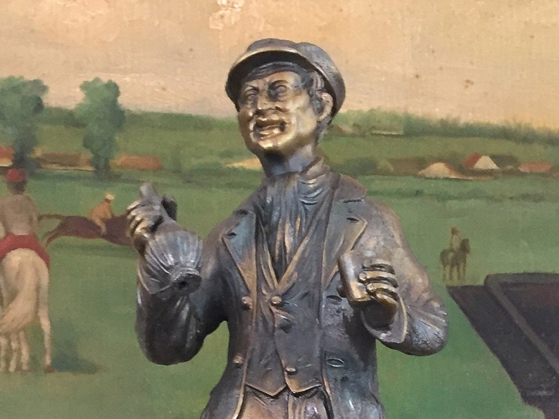 May include: A bronze statue of a man in a cap and coat, holding a bottle and a cup, with a painted backdrop of a horse race. The statue has a detailed, aged appearance, capturing a moment of excitement.