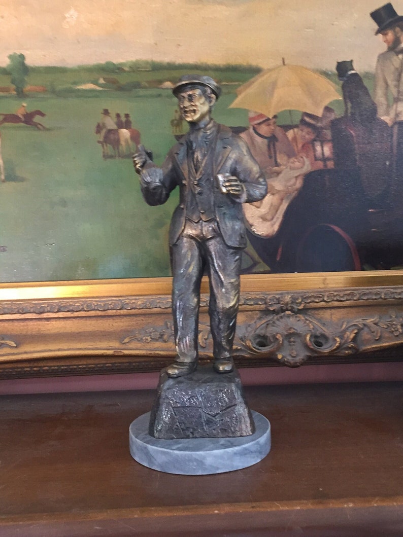May include: A bronze-colored statue of a man in a suit and cap, holding a glass and gesturing. The statue is mounted on a stone base with a circular marble base. The background features a framed painting with a horse racing scene.