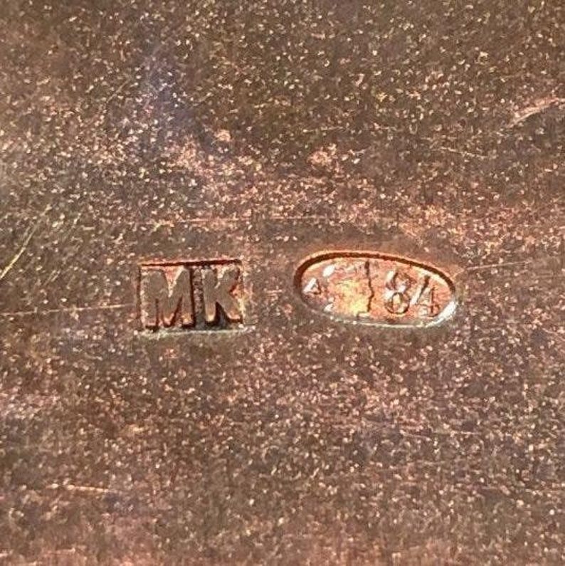Op de afbeelding: Close-up van een metalen oppervlak met twee markeringen. De eerste markering is een vierkant met de letters "MK" erin. De tweede markering is een ovaal met de cijfers "84" erin.