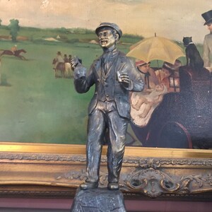 May include: A bronze-colored statue of a man in a suit and cap, holding a small object in each hand. The statue is set against a backdrop of a framed painting depicting a horse race and people in a carriage.