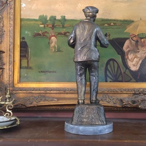 May include: A bronze-toned statue of a man in a cap and jacket, standing on a textured base atop a marble-like circular platform. The statue is positioned in front of a framed painting depicting a horse race scene.
