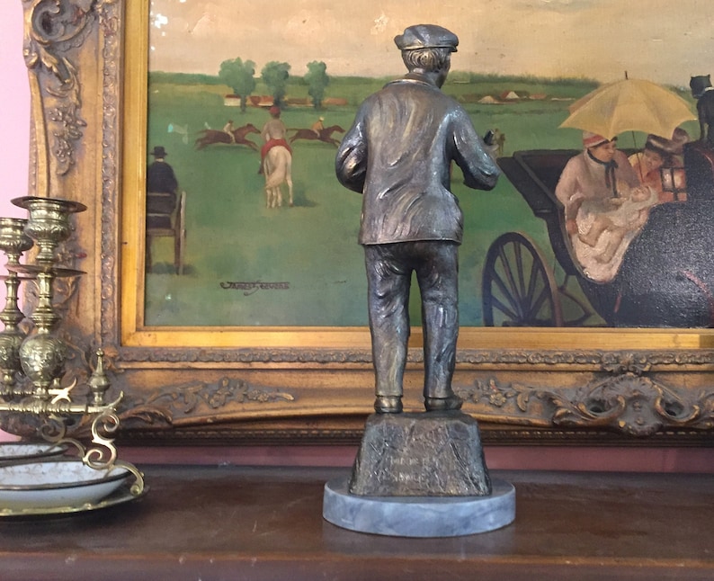 May include: A bronze-toned statue of a man in a cap and jacket, standing on a textured base atop a marble-like circular platform. The statue is positioned in front of a framed painting depicting a horse race scene.
