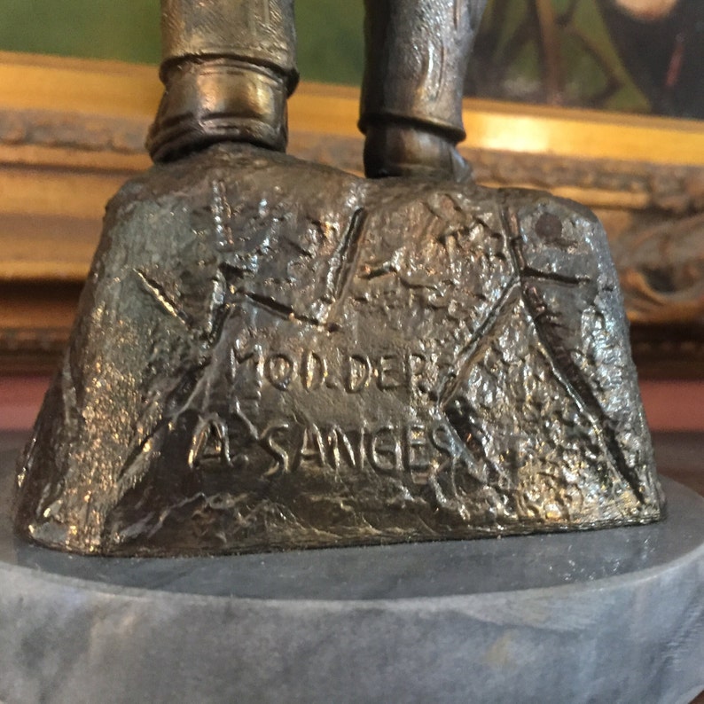 May include: Bronze sculpture base with the inscription "MOD. DERI A. SANGES" on a textured surface. The sculpture is mounted on a gray marble base. The legs of the sculpture are visible above the base.