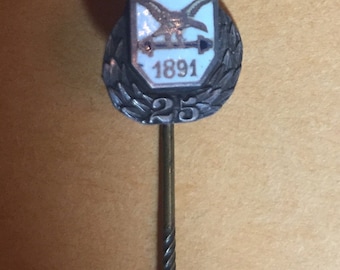 Op de afbeelding: Een zilveren broche met een wit geëmailleerd schild waarop een vogel met uitgespreide vleugels en een pijl te zien is. Het schild heeft de tekst "A.S.V. 1891" en een krans met het nummer "25" aan de onderkant.