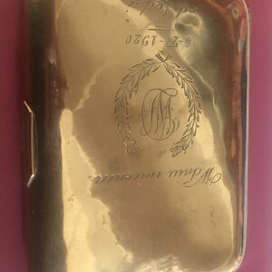 Op de afbeelding: Een gouden metalen sigarettenkoker met een gegraveerd monogram en de inscriptie "0367-L7-9".