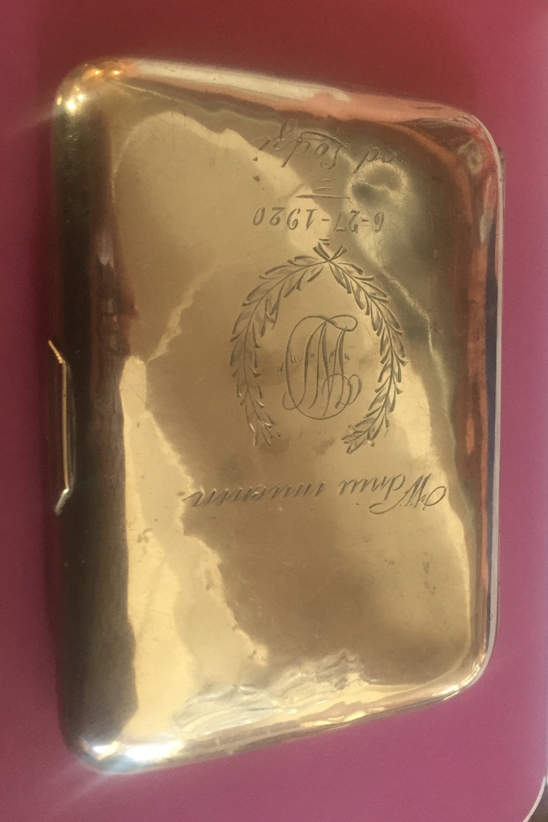 Op de afbeelding: Een gouden metalen sigarettenkoker met een gegraveerd monogram en de inscriptie "0367-L7-9".