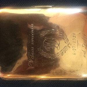 Op de afbeelding: Een gouden metalen sigarettenkoker met een gegraveerd ontwerp. Het ontwerp omvat een krans, een monogram en de tekst "6-27-12 - 9-0826".