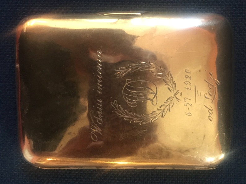 Op de afbeelding: Een gouden metalen sigarettenkoker met een gegraveerd ontwerp. Het ontwerp omvat een krans, een monogram en de tekst "6-27-12 - 9-0826".