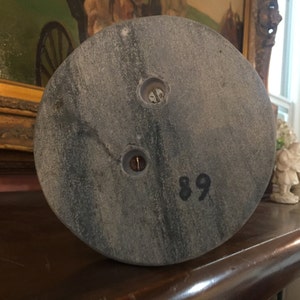 May include: A circular, gray stone object with two screw holes and the number 89 printed in black. The stone has a mottled appearance and is likely part of a larger structure or decorative item.