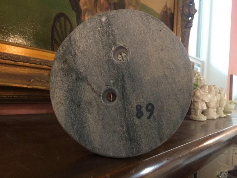 May include: A circular, gray stone object with two screw holes and the number 89 printed in black. The stone has a mottled appearance and is likely part of a larger structure or decorative item.
