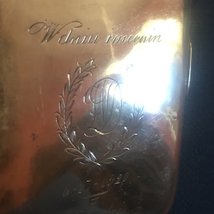Op de afbeelding: Een gouden metalen doos met een gegraveerd opschrift dat "W dniu imienin" luidt en een krans met de letter "D" in het midden.