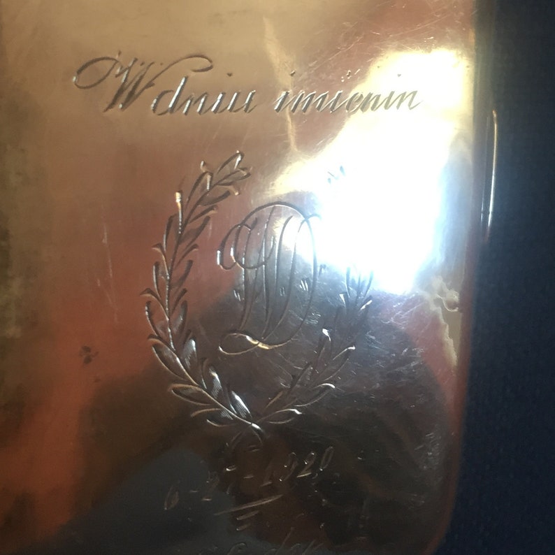 Op de afbeelding: Een gouden metalen doos met een gegraveerd opschrift dat "W dniu imienin" luidt en een krans met de letter "D" in het midden.