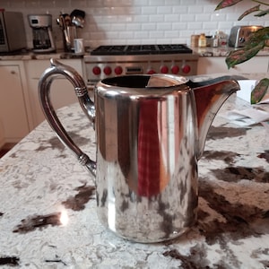 May include: A silver pitcher with a handle and a spout. The pitcher is sitting on a white countertop.