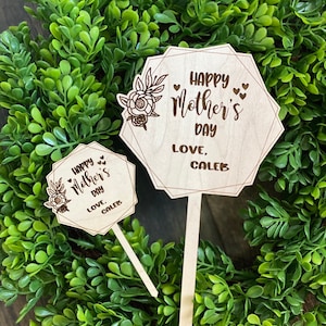May include: Two wooden signs with a geometric shape and a floral design. The larger sign says "Happy Mother's Day Love, Caleb". The smaller sign says "Happy Mother's Day Love, Caleb".