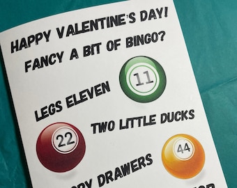 Puede incluir: Una tarjeta de San Valentín con temática de bingo. La tarjeta tiene una bola de bingo verde, roja, azul y naranja con los números 11, 22, 69 y 44. El texto de la tarjeta dice "¡Feliz Día de San Valentín! ¿Te apetece un poco de bingo?"  "Legs Eleven", "Two Little Ducks", "Droopy Drawers", y "A Meal for Two with a Hairy View".