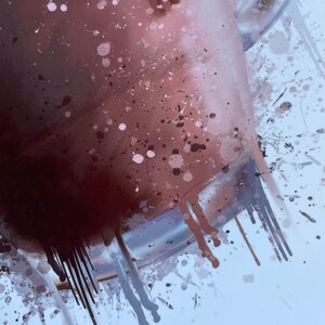 Puede incluir: Primer plano de una taza de vidrio llena de un l&iacute;quido marr&oacute;n oscuro, con salpicaduras de pintura marr&oacute;n y blanca sobre un fondo blanco.
