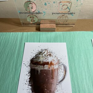 Puede incluir: Una ilustraci&oacute;n de estilo acuarela de una bebida de chocolate caliente con crema batida y un chorrito de chocolate. La bebida est&aacute; en una taza de vidrio transparente con asa. El fondo es blanco con salpicaduras de pintura gris.