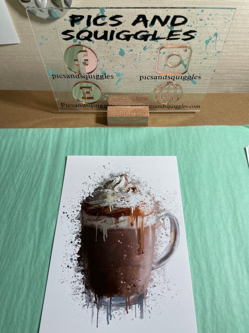 Puede incluir: Una ilustraci&oacute;n de estilo acuarela de una bebida de chocolate caliente con crema batida y un chorrito de chocolate. La bebida est&aacute; en una taza de vidrio transparente con asa. El fondo es blanco con salpicaduras de pintura gris.