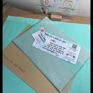 Puede incluir: Un paquete de papel azul claro y marr&oacute;n con el texto "Please do not bend" en el papel marr&oacute;n. Una tarjeta peque&ntilde;a con el texto "Thank you so much for your order" y "Pics and Squiggles" es visible en la parte superior del paquete.