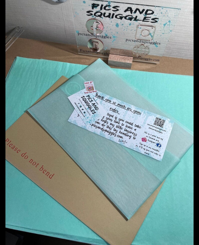 Puede incluir: Un paquete de papel azul claro y marr&oacute;n con el texto "Please do not bend" en el papel marr&oacute;n. Una tarjeta peque&ntilde;a con el texto "Thank you so much for your order" y "Pics and Squiggles" es visible en la parte superior del paquete.