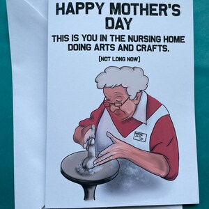May include: A humorous Mother's Day card featuring a cartoon illustration of an older woman in a nursing home, wearing a red shirt and a white apron, working on a pottery wheel. The text reads "Happy Mother's Day. This is you in the nursing home doing arts and crafts. [Not long now]"
